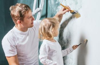Image of a paint brush being used to apply paint to a wall. The brush is held in the hand of a man who is painting a wall with his young daughter.The man and daughter are applying the paint to the wall in a smooth, even coat.
